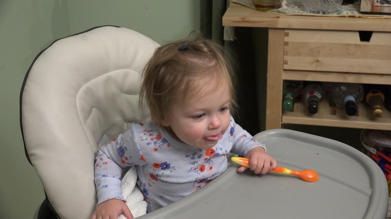 The toddler remains in the high chair in the Westminster home interior, leaning forward with one hand on the tray and the orange spoon near the mouth.