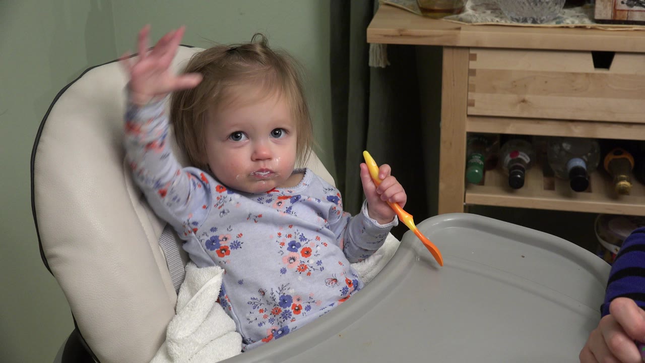 At 0:01 in Westminster, Colorado, the child faces the camera from the booster seat, with one hand raised and the yellow and orange spoon held over the tray.