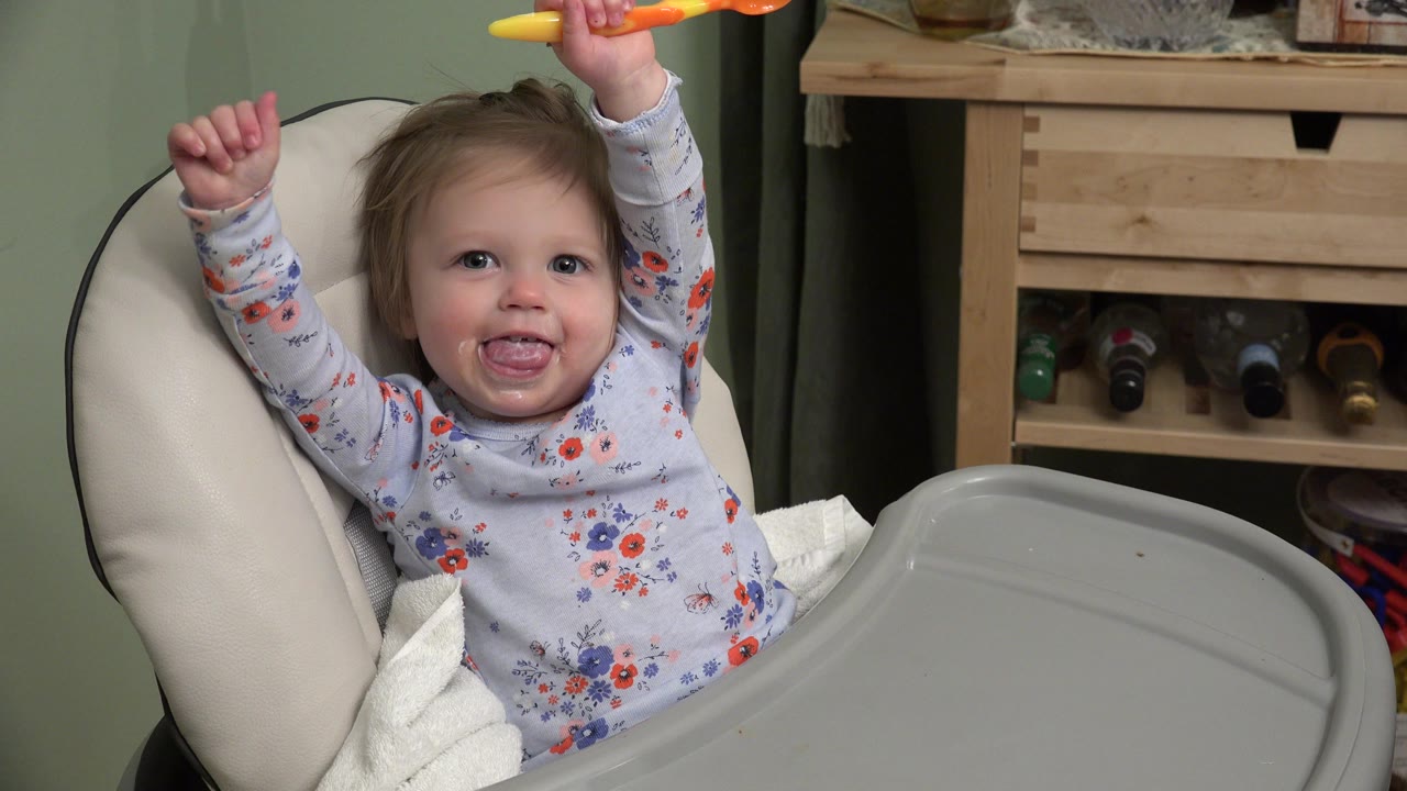 Baby in a high chair raises an orange spoon and smiles.