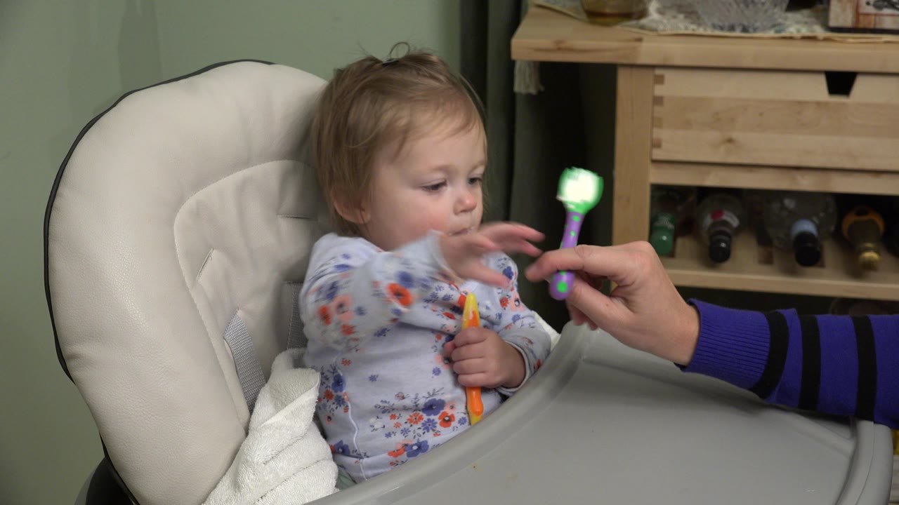 The toddler remains seated in the booster chair, looking at the green spoon extended by an adult hand beside the gray tray in the Westminster home interior.