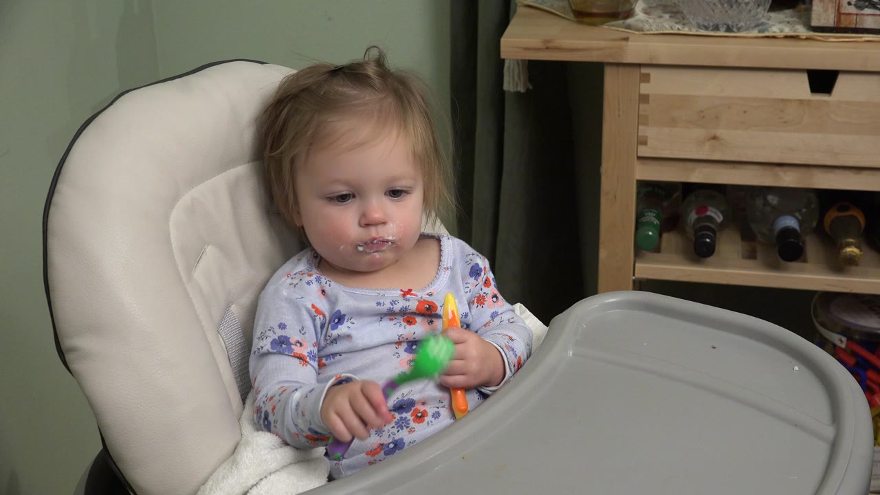 Infant seated in a white highchair holding colorful utensils.