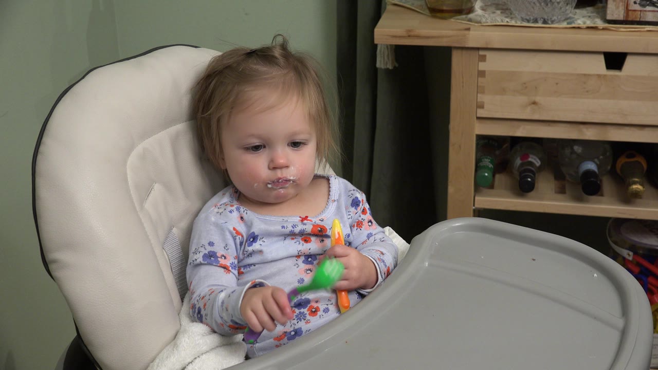 In Westminster, Colorado, the infant looks toward the tray while seated in the white highchair, with food residue visible around the mouth and colorful utensils in hand.