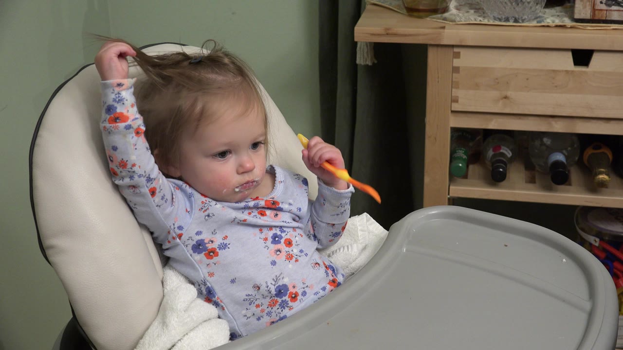 Baby in a high chair holding an orange spoon near the mouth.