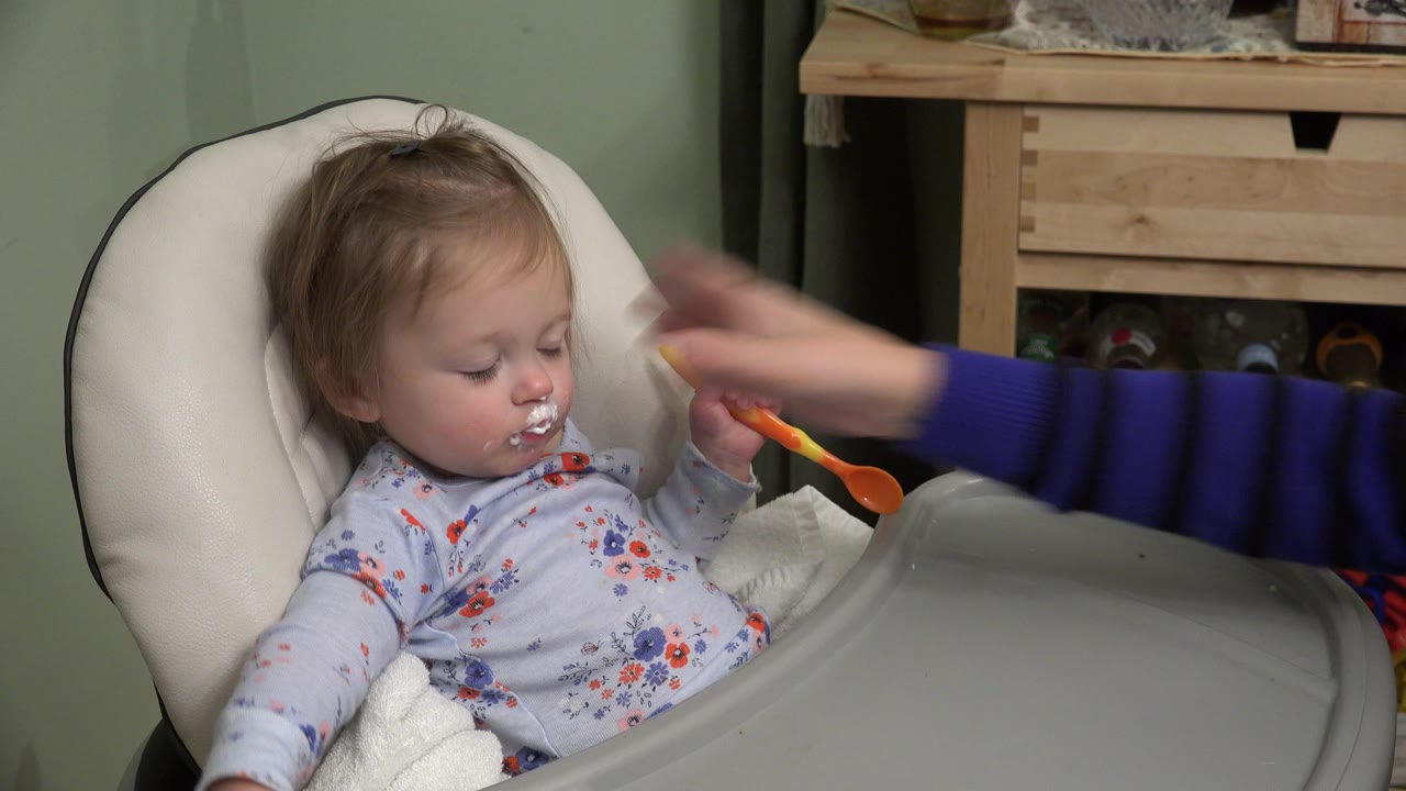 Toddler in high chair reaching near an adult hand and spoon.
