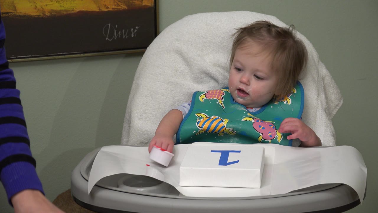 In Westminster, Colorado, the baby remains centered in the high chair, looking down toward the red paint cup and white paper with a blue painted shape.