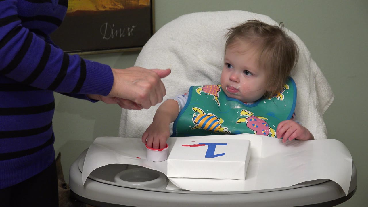 In Westminster, Colorado, a toddler in a white high chair looks toward an adult's hand giving a thumbs-up beside paint paper on the tray.