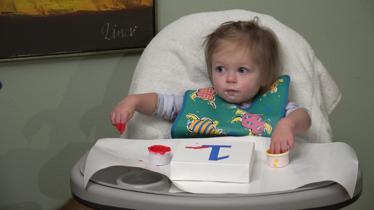 Baby in a high chair holding red paint at a tray.