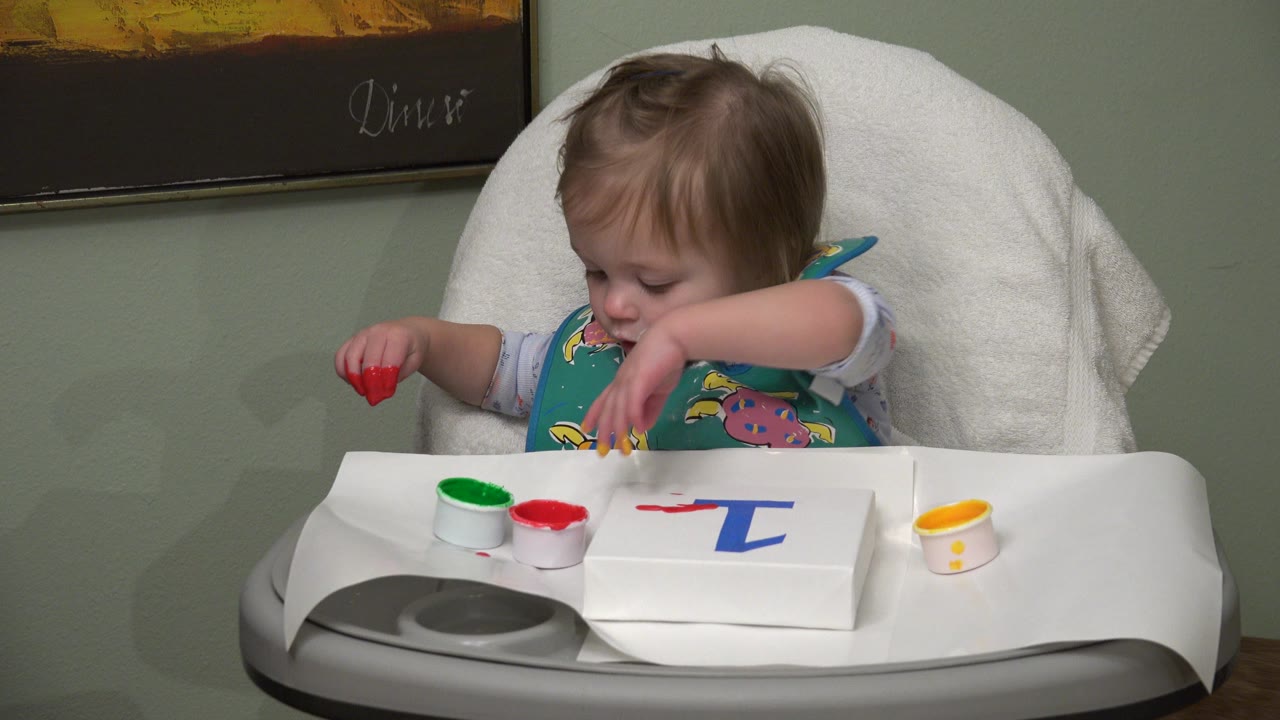 The Westminster toddler looks down at the tray while holding red paint in one hand and moving the other hand near the paint cups and canvas.
