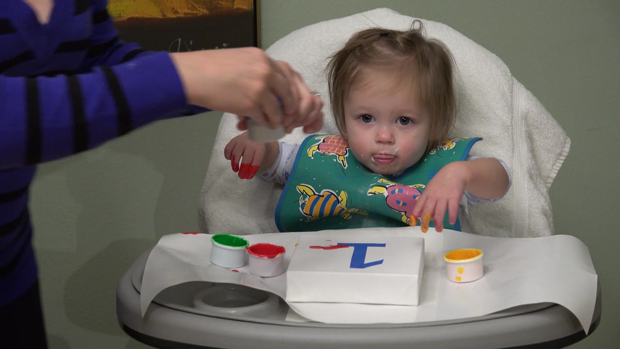 Toddler in a highchair with paint cups on the tray as an adult hand reaches in.