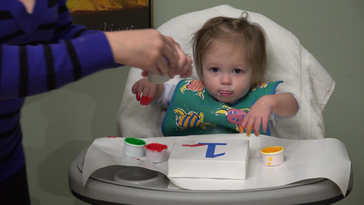 In Westminster, Colorado, the toddler looks toward the camera from the highchair as an adult hand remains near the paint cups and paper on the tray.