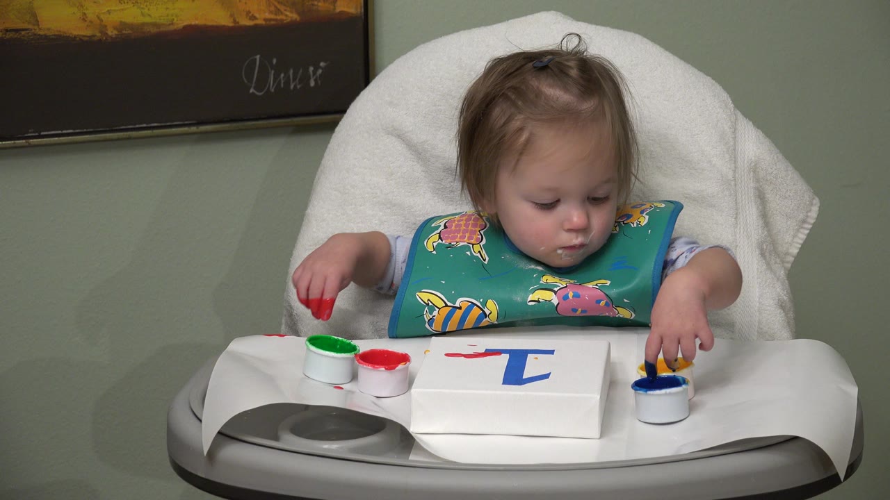 Blonde baby in a high chair reaching toward paint cups.
