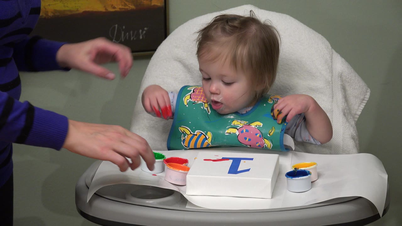 Baby in a high chair reaches toward red paint on a tray.
