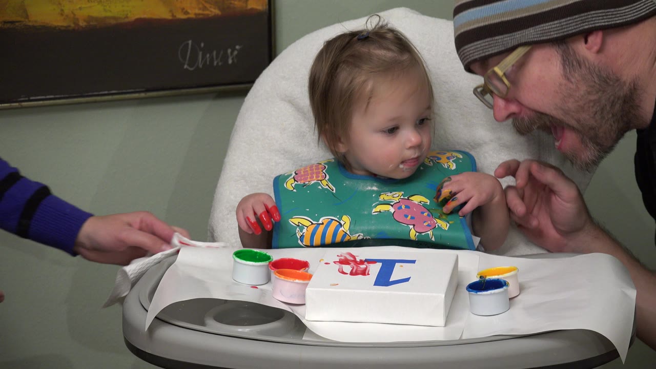 Toddler in high chair with paint cups and parent leaning in.