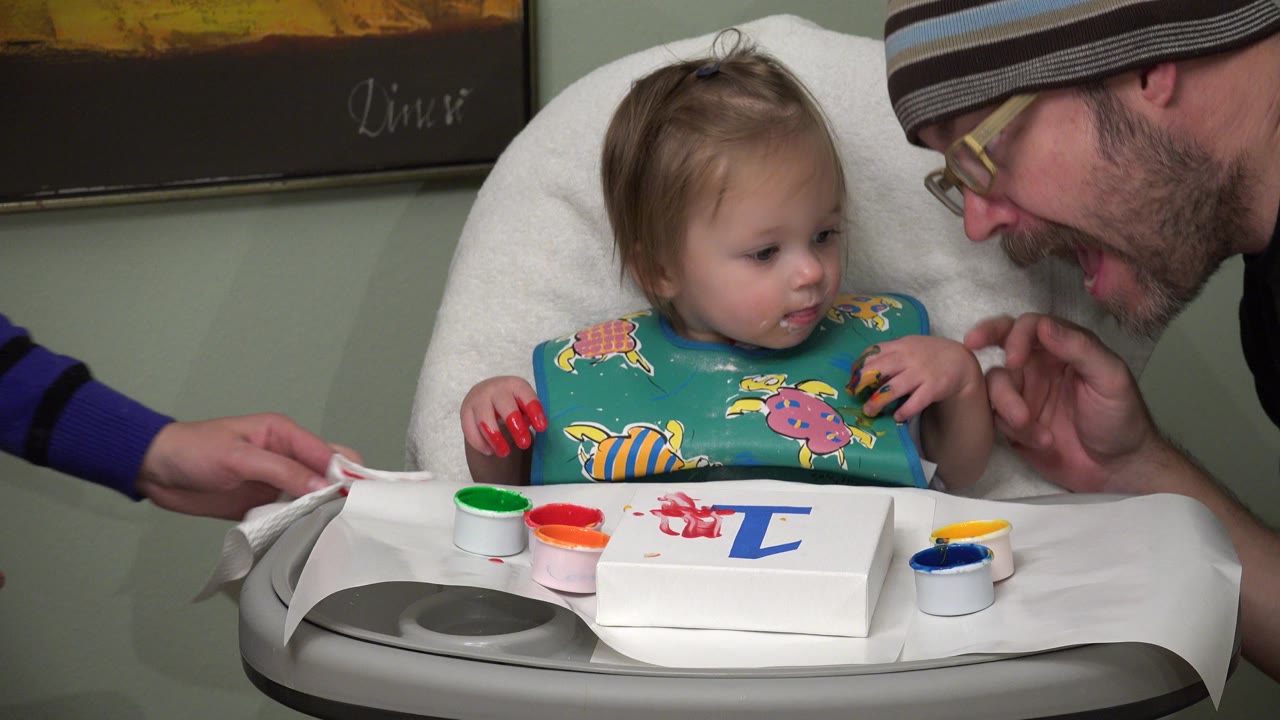 In Westminster, Colorado, the toddler looks toward an adult leaning near the high chair while small open paint cups and a white sheet with a blue letter sit on the tray.