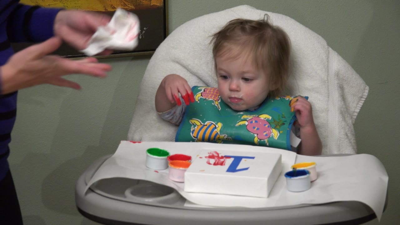 Toddler girl seated at a tray with paint cups as an adult holds a wipe nearby.