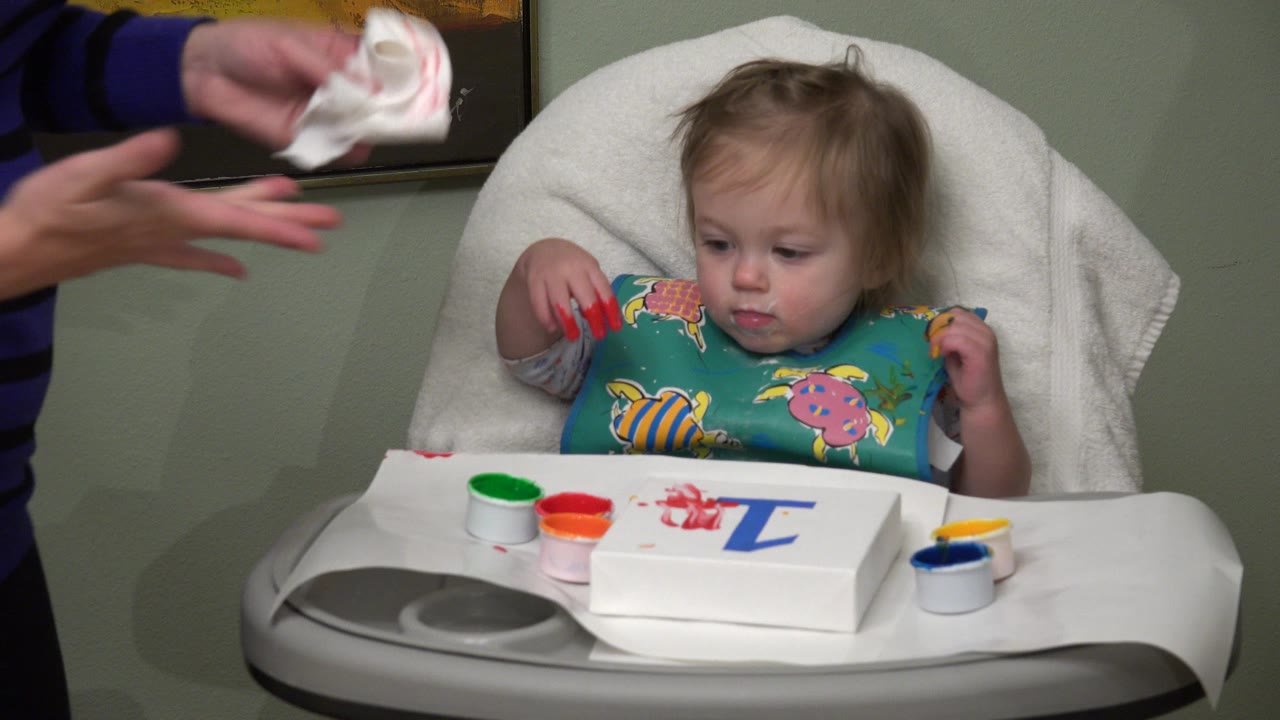The toddler remains centered in the child seat with red paint on her fingers, open paint containers on the tray, and an adult hand with a wipe at frame left.