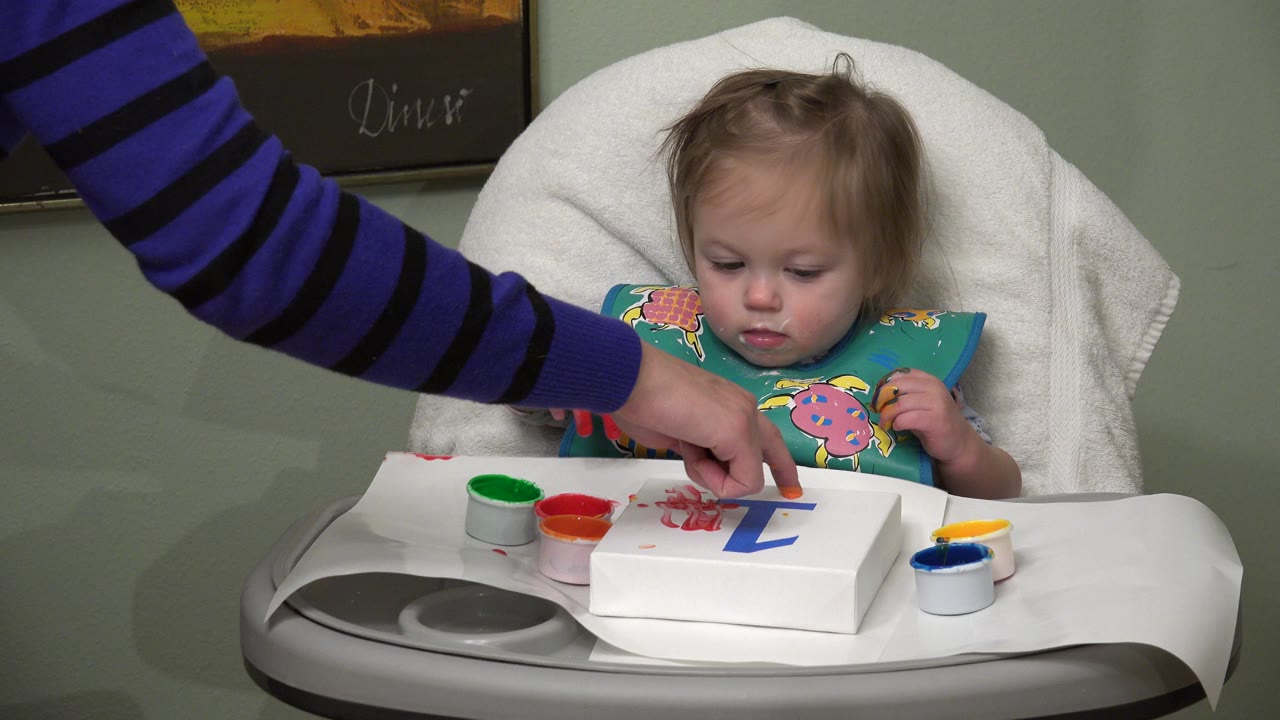 Baby in a high chair while an adult points at paint on paper.