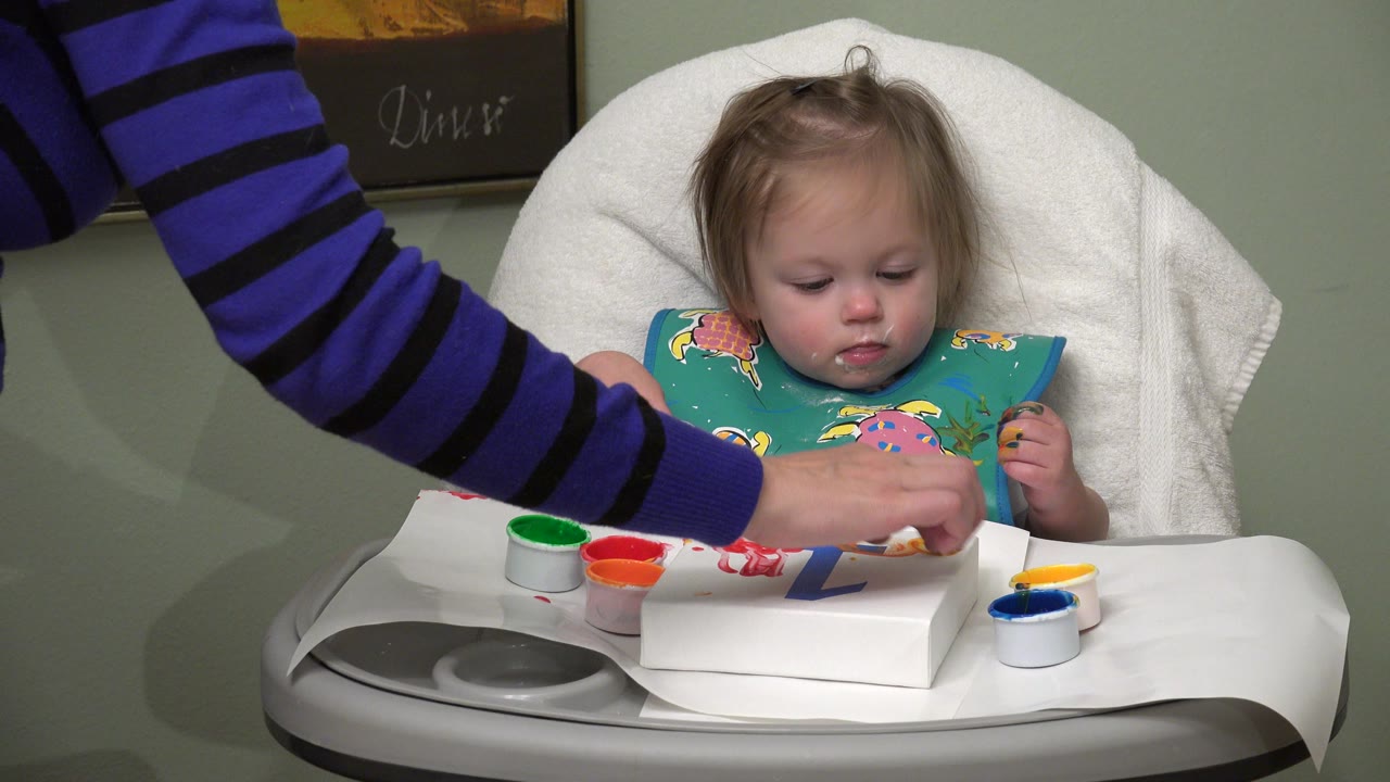 The child remains centered in the high chair while an adult arm reaches in from the left over red, green, yellow, and blue paint containers.