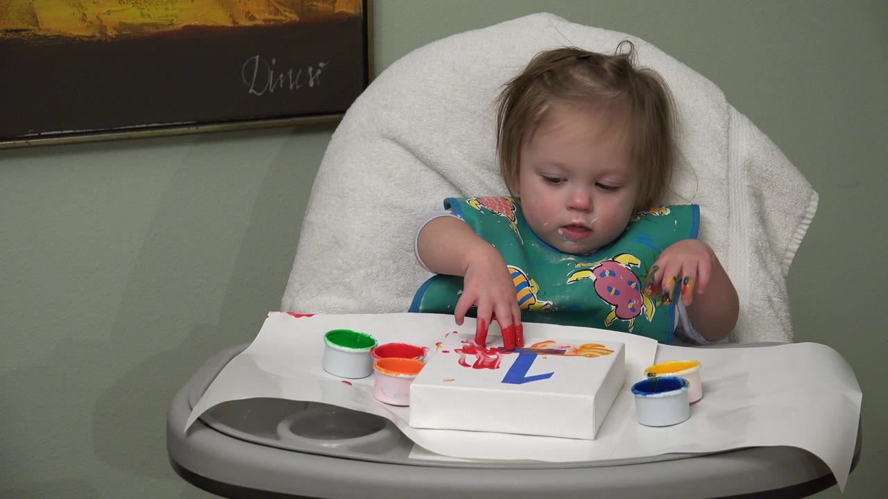 Blonde toddler in a high chair touches red paint on a white canvas.