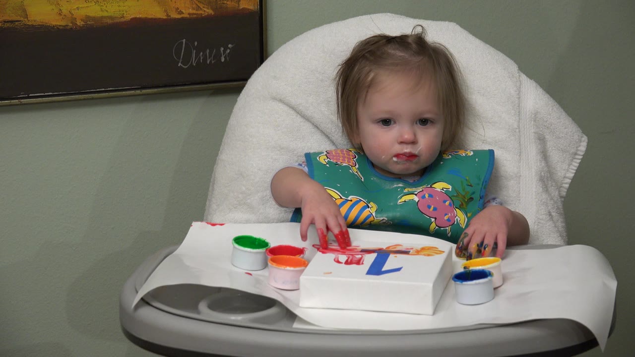 Blonde toddler in a high chair touches red paint on paper.