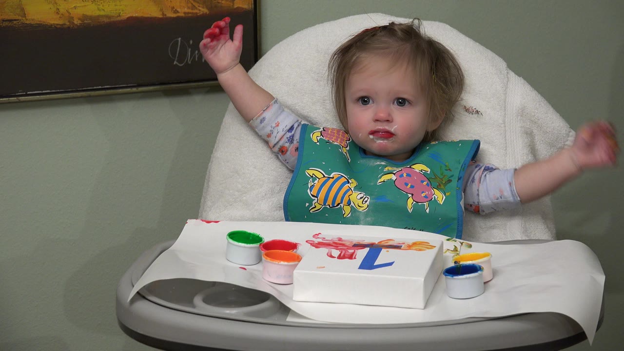 The baby faces forward in the highchair with one red-painted hand lifted, the other arm extended, and finger paint supplies spread across the tray in Westminster, Colorado.