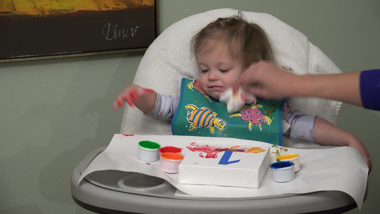 In Westminster, Colorado, a baby wearing a colorful bib sits in a white high chair while an adult hand wipes the baby's mouth with a napkin above paint cups on the tray.
