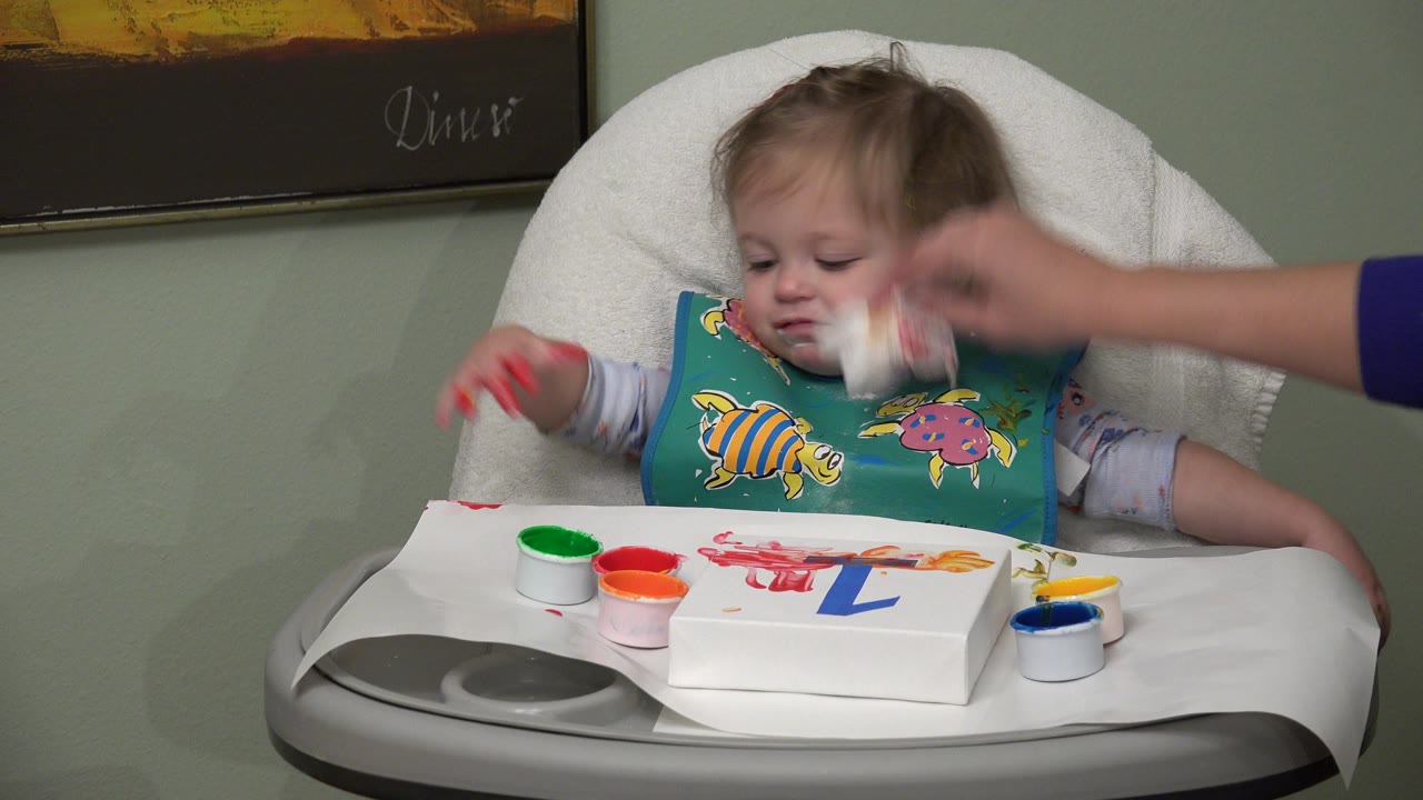In Westminster, Colorado, the baby looks down in the high chair as the adult hand continues wiping near the mouth, with red, green, orange, yellow, and blue paint cups on the tray.