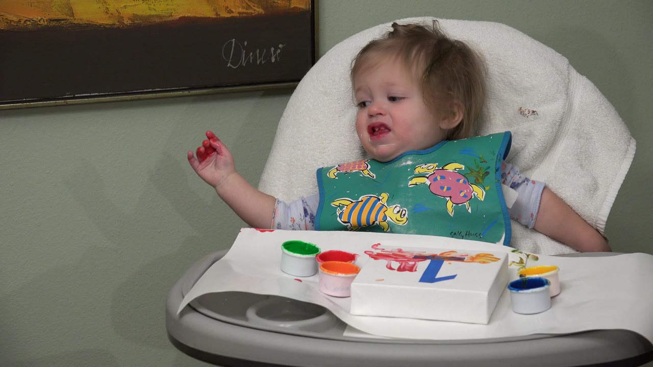 Toddler in Westminster, Colorado looks off to the side with red paint on raised fingers while paint cups and a number activity remain on the high-chair tray.