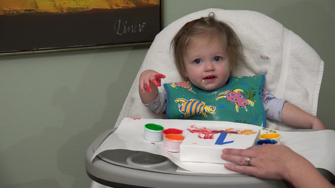 At 0:00, a baby in Westminster, Colorado sits in a white high chair with red paint on one hand, paint cups on the tray, and a board showing a blue number one.