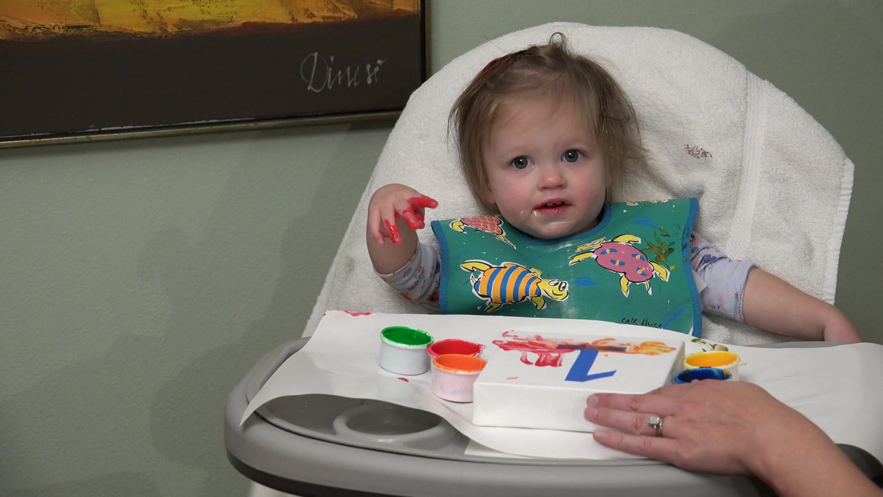 At 0:01, the baby remains centered in the high chair while an adult hand steadies the activity board with the blue number one and small paint cups arranged along the tray.