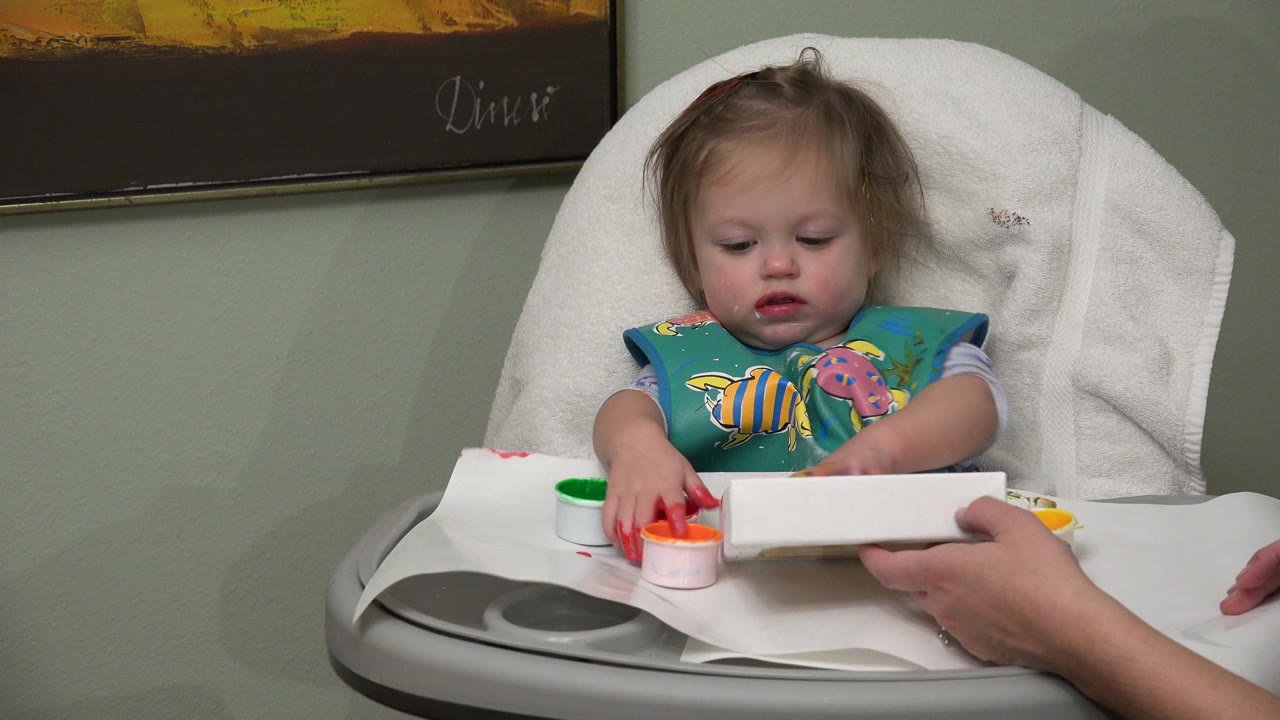 Toddler in a highchair reaches toward paint cups and a white card.
