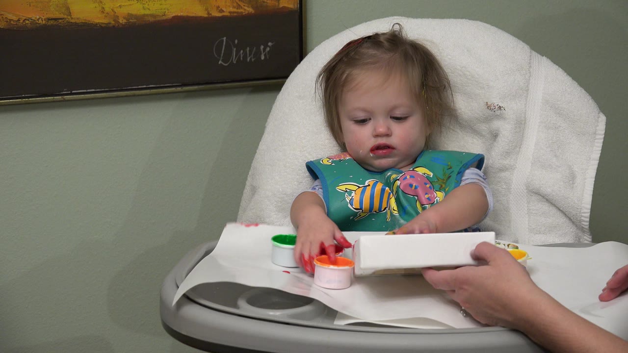 In Westminster, Colorado, the child remains centered in the highchair as red paint sits near their fingers and the adult steadies the white card.