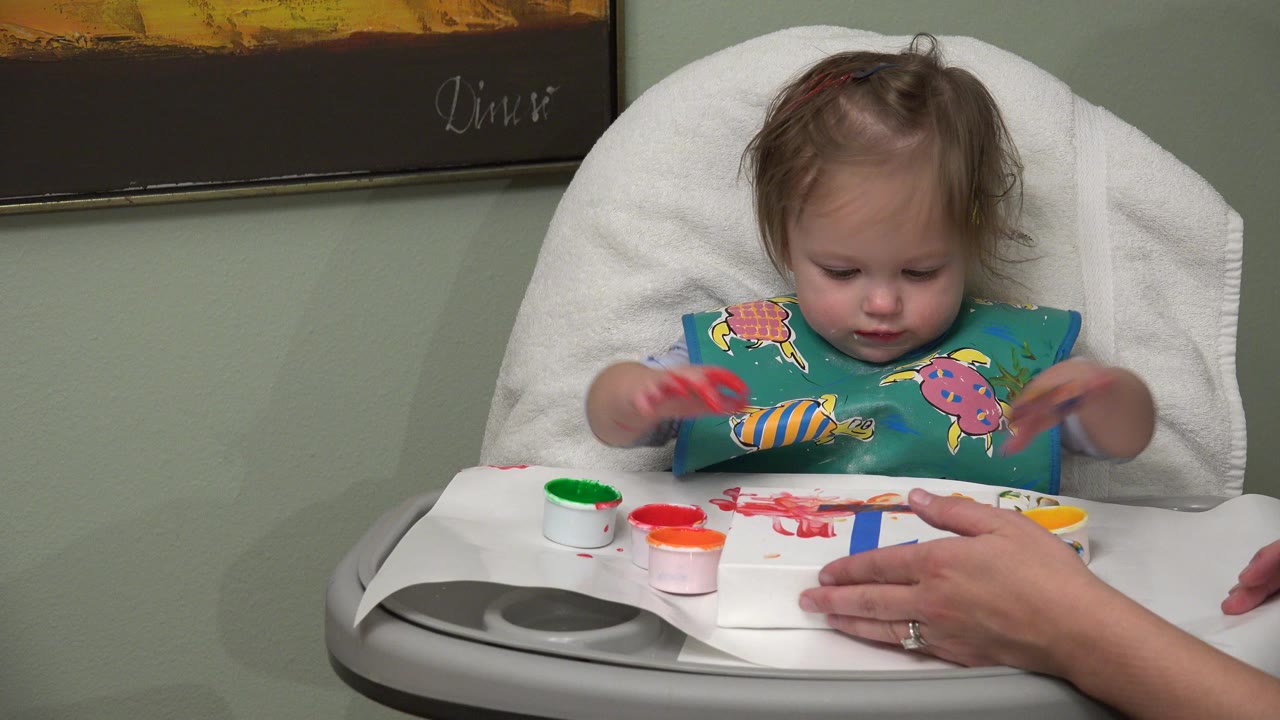 In Westminster, Colorado, a baby girl in a high chair looks down while touching red paint on paper, with small paint cups and an adult hand on the tray.