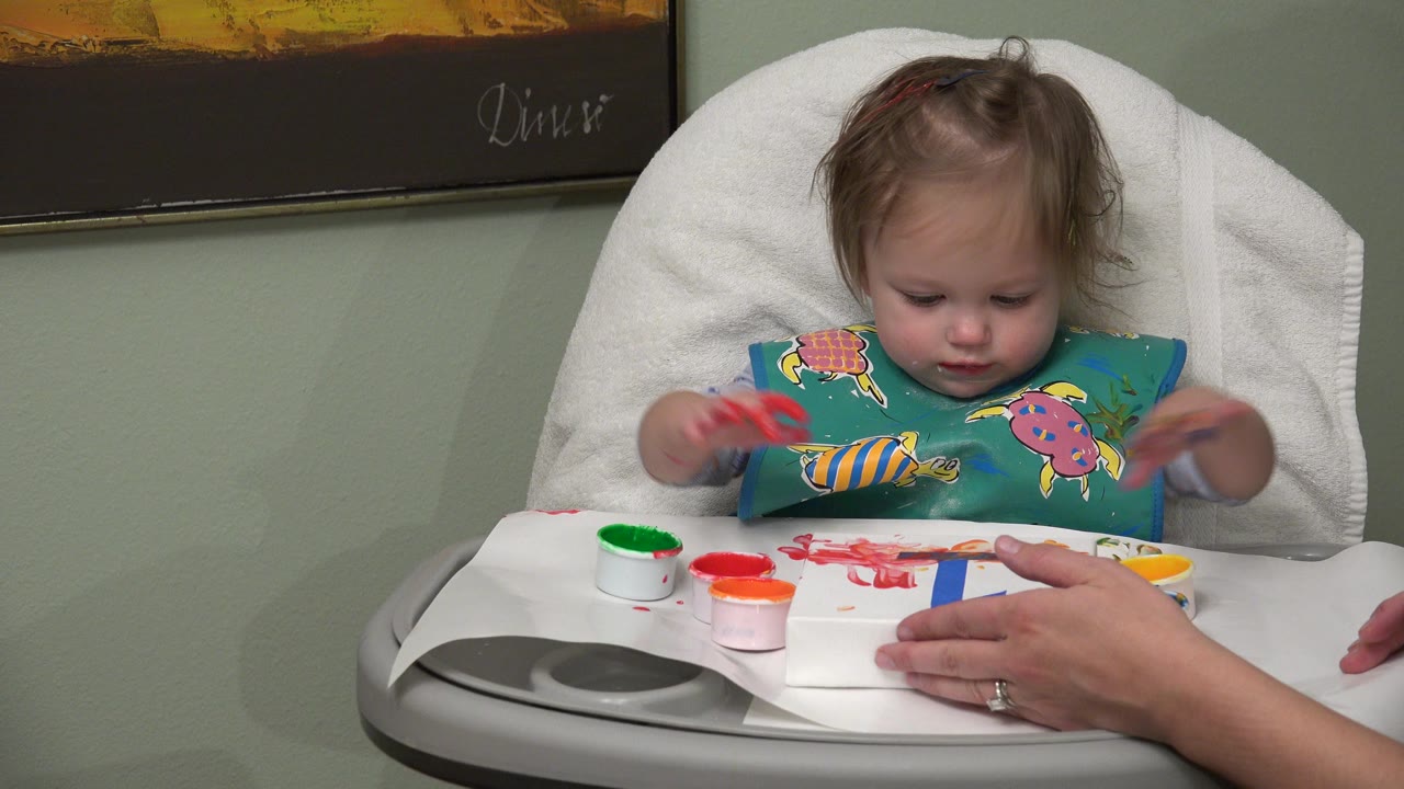 In Westminster, Colorado, the baby girl keeps her gaze on the paint-covered paper as an adult hand holds the sheet steady on the high chair tray.