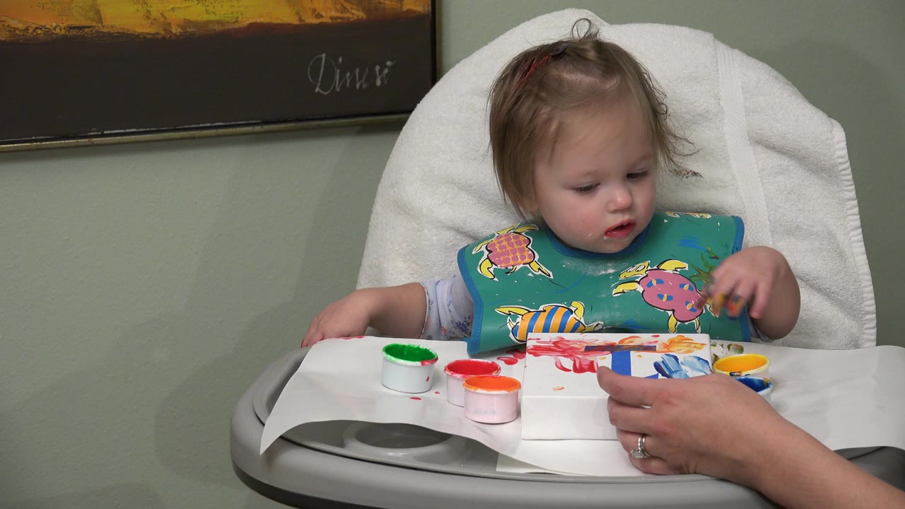 Toddler in a high chair painting at a tray with adult hands nearby.