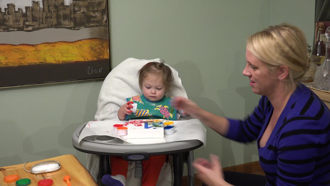 Baby in a highchair with paint while a woman reaches toward the tray.