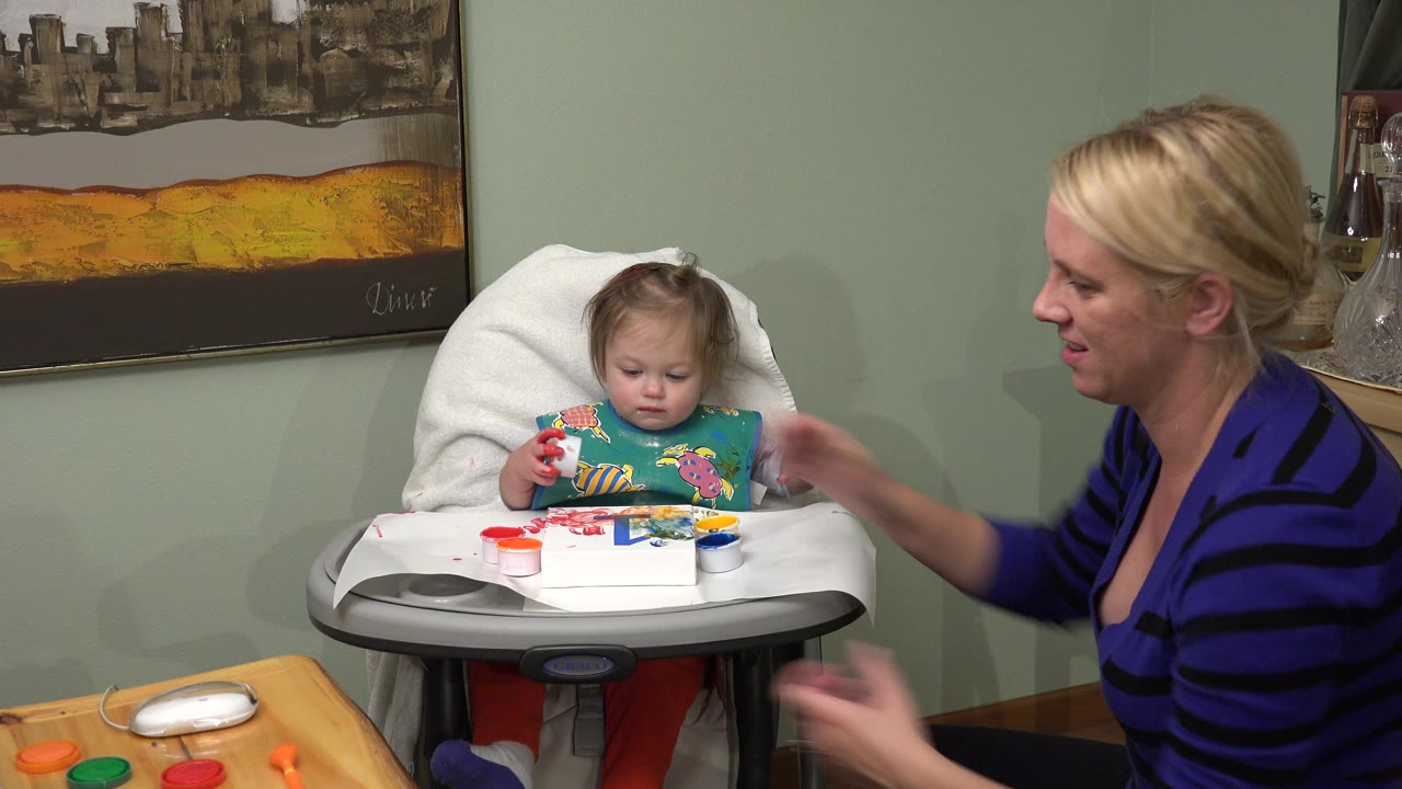 In Westminster, Colorado, the baby looks down at the paint tray while the woman sits close on the right, with wall art and a green interior wall behind them.