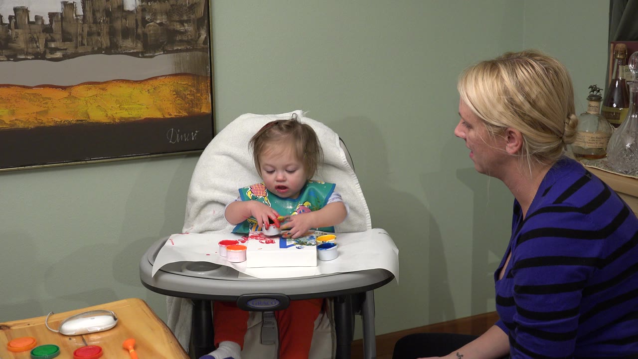Toddler in a high chair plays with colorful items while a woman sits beside her.