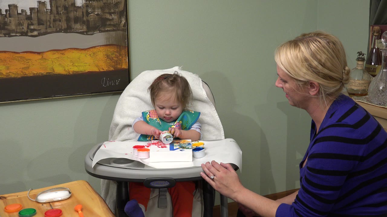 Toddler in a high chair playing while a woman sits beside her.