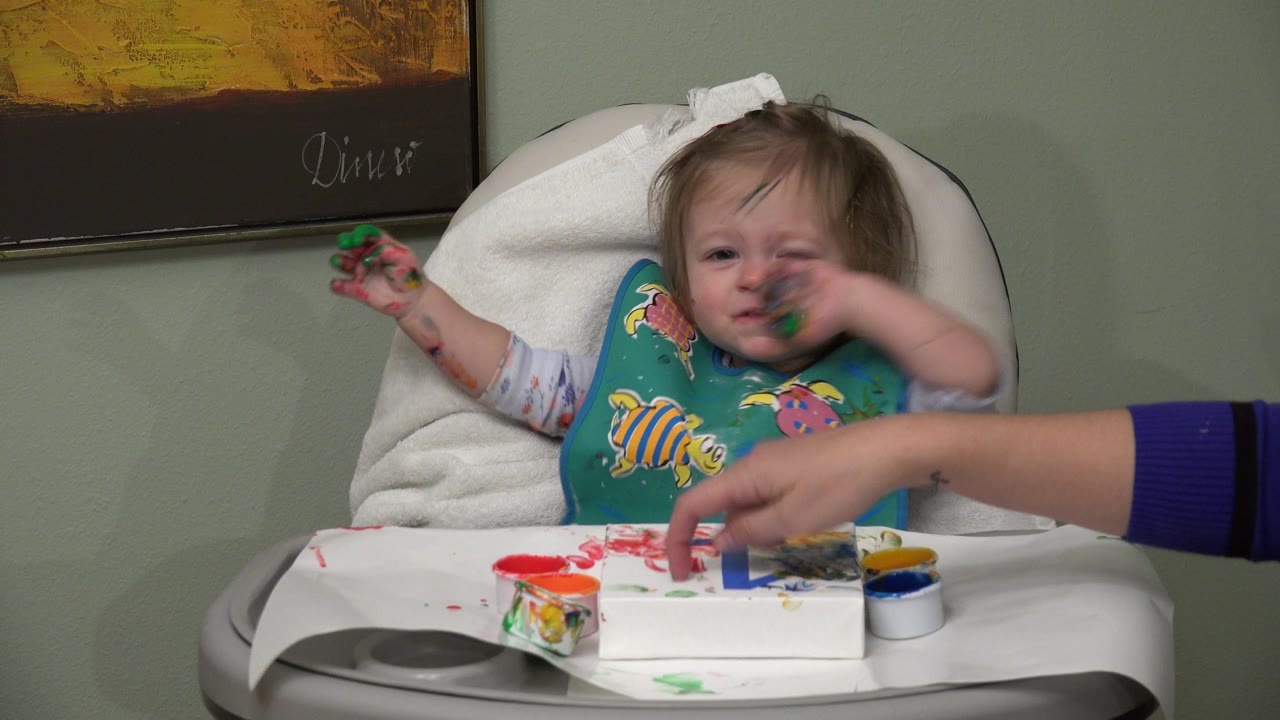 In Westminster, Colorado, an adult hand reaches across the high-chair tray as the baby raises green-painted fingers near her face with paint supplies spread in front of her.