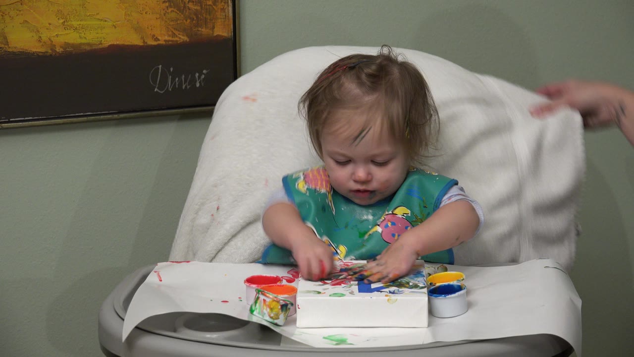 At 0:00 in Westminster, Colorado, a toddler in a high chair presses fingers into paint on a white tray while an adult hand enters from the right near a towel-covered chair back.