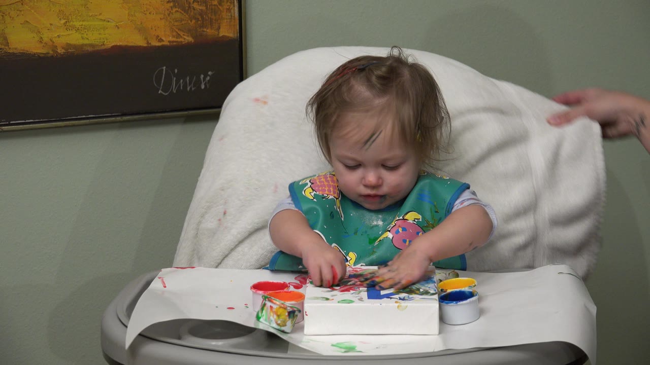 At 0:01 in Westminster, Colorado, the toddler looks down at paint-smeared paper and paint pots while an adult hand remains partly visible at the right edge.