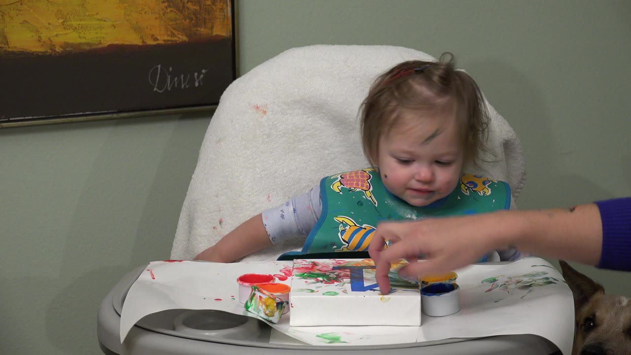 The child looks down at the painted tray as an adult hand points or presses near a yellow paint area in the Westminster home interior.