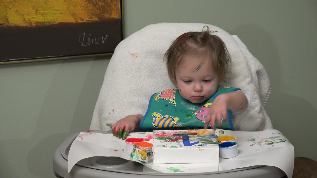 Baby in a highchair reaches toward finger paint on a tray.