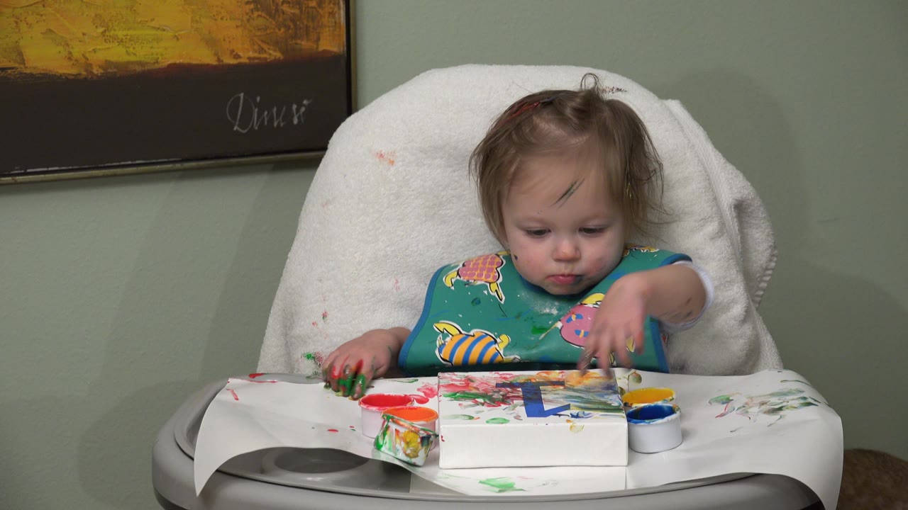The baby keeps one hand near the left side of the tray and extends the other over the painted paper, with red, green, blue, yellow, and orange paint visible on the highchair surface.