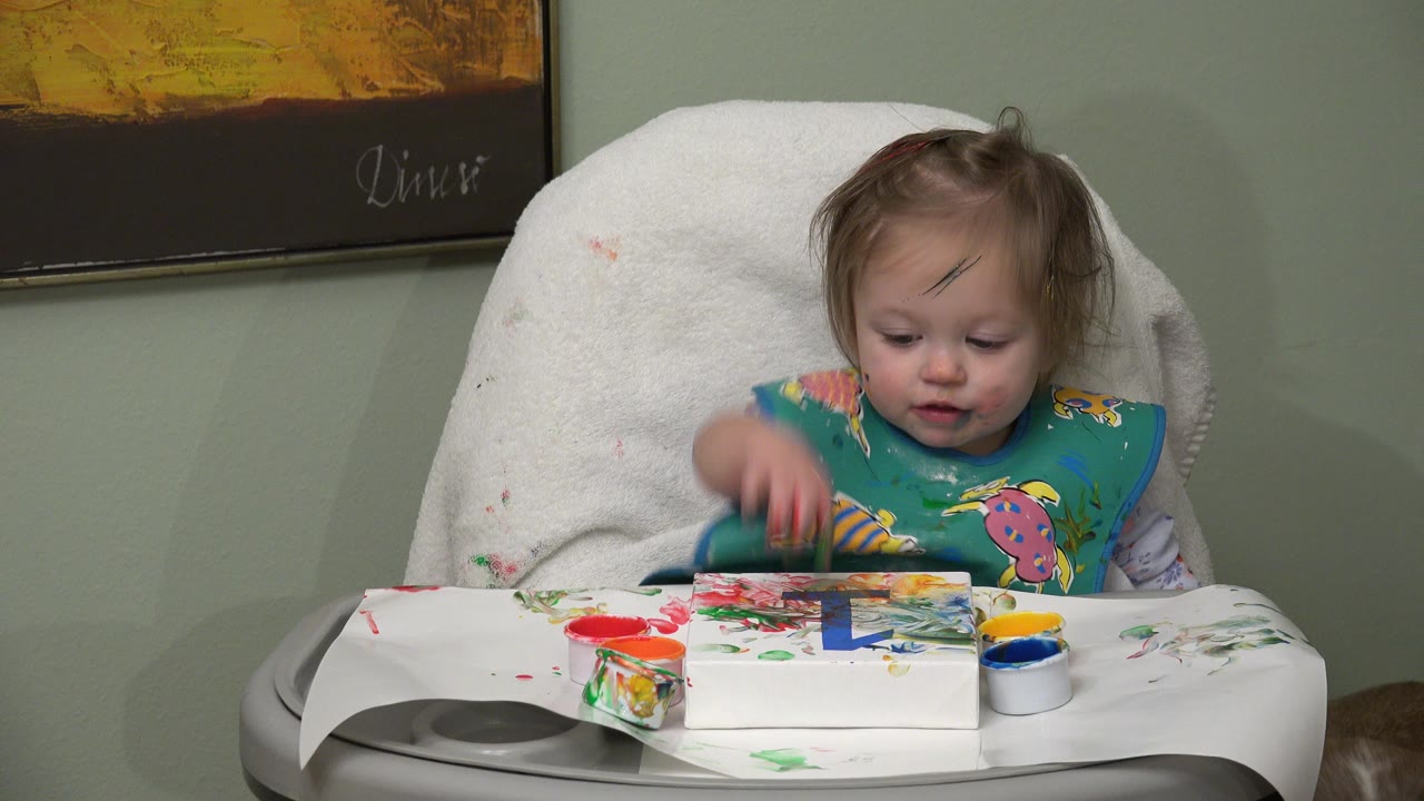 The Westminster toddler raises a paint-marked hand above the tray while red, green, blue, and yellow paint marks spread across the white surface.
