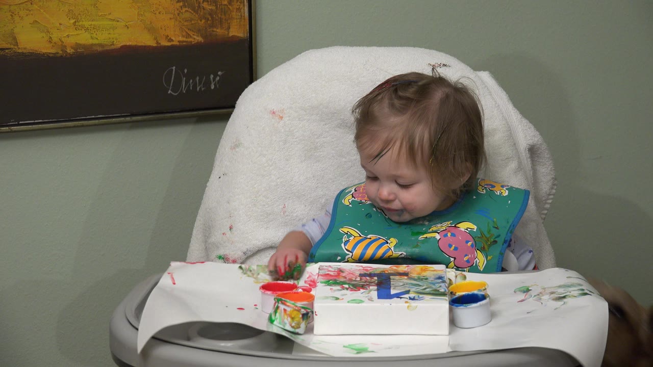 An infant in Westminster, Colorado sits in a high chair with a colorful bib, touching red and green paint on a tray with cups and paper in front.