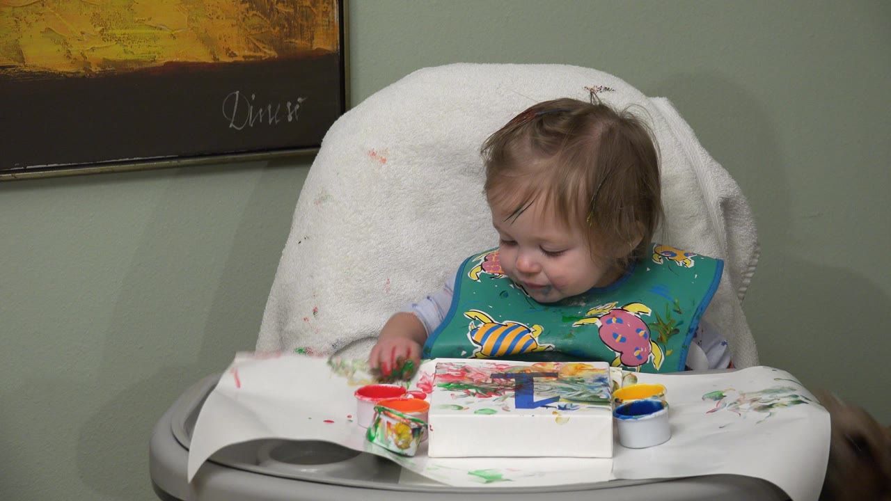 The Westminster, Colorado infant looks down at the paint-covered tray, with red and blue paint cups and a marked paper surface in front of the high chair.