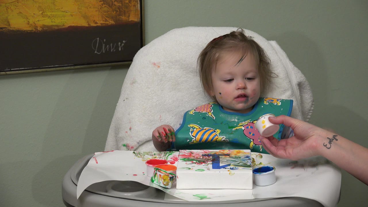 Baby in a high chair with finger paint and an adult hand holding a small white cup.