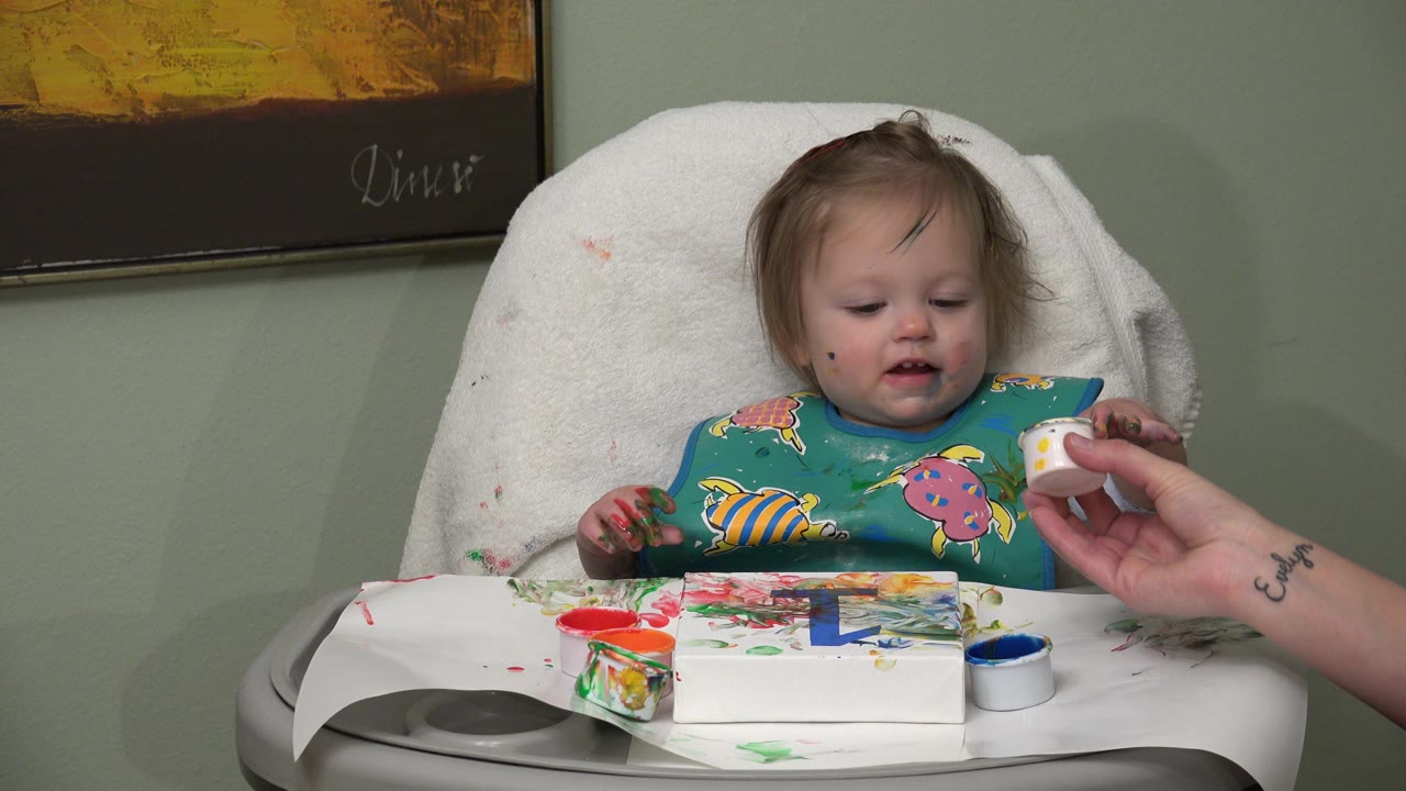 In Westminster, Colorado, the baby leans slightly toward the adult hand and small white cup, with blue, green, red, and yellow paint visible on the tray and box.